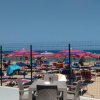 Spiaggia affollata con ombrelloni rosa e sedie, vista da recinto.