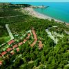 Spiaggia con mare turchese, pineta e complesso di case con tetti rossi in lontananza.
