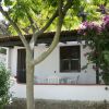 Una piccola casa bianca con terrazza, tavolo rotondo e sedie. Albero e fiori viola accanto.