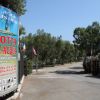 Entrata di un villaggio turistico con cartello informativo e vegetazione.