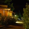 Strada esterna di notte con piante e illuminazione su edifici arancioni.