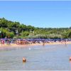 Spiaggia affollata con ombrelloni blu e vegetazione collinare.