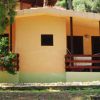 Bungalow giallo con terrazza in legno, circondato da alberi.