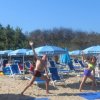 Persone che fanno esercizi di stretching su una spiaggia con sdraio blu e ombrelloni.